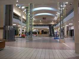 The east court of the Crossroads Mall, facing the anchors. All storefronts shown are empty, with the elevator tower to the left of the image, with kids rides, a play area, and escalator in the middle of the court.