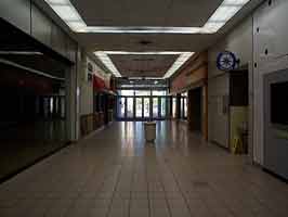 The south entrance corridor of the Crossroads Mall, looking towards the door. To the left and right are empty storefronts, with a trashcan in the middle.