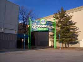 The south entrance of the Crossroads Mall. In front is a sign for the mall on blue mesh, supported by lime metal posts. Two trees are on either side of the entrance sidewalk.