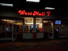 The West Mall 7 theater. In the middle is a ticket booth, lit by many incadescant bulbs. Above is a fancy typeface sign reading 'WestMall 7', and to the left and right are glass doors.