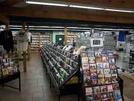A CD rack in the 41st street Last Stop, containing hundreds of various CDs. DVDs line the far wall, with a display stand of DVDs in front of the rack. To the right of the rack are video games and accessories.